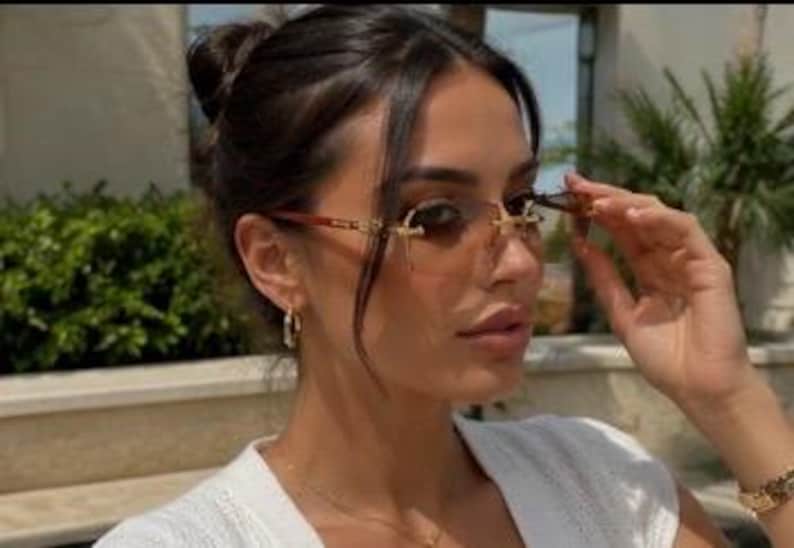 May include: Rimless sunglasses featuring rectangular gradient brown lenses and thin gold-tone metal temples with brown acetate tips. The bridge and hinges are crafted from polished gold-tone metal, providing a sleek and lightweight appearance.