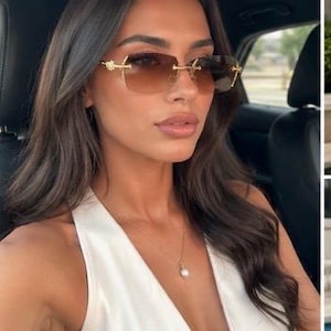May include: Rimless sunglasses featuring rectangular brown gradient lenses and gold-toned metal temples with ornate detailing at the hinges. The eyewear is paired with a delicate gold chain necklace featuring a single white pearl pendant.