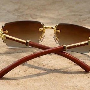 May include: Rimless sunglasses featuring gradient brown lenses that transition from dark to light. The frame includes gold-toned metal hinges with intricate engravings and smooth, dark reddish-brown wooden temples that curve inward at the ends.