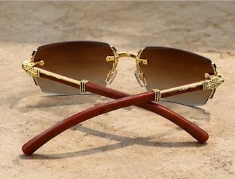 May include: Rimless sunglasses featuring gradient brown lenses that transition from dark to light. The frame includes gold-toned metal hinges with intricate engravings and smooth, dark reddish-brown wooden temples that curve inward at the ends.