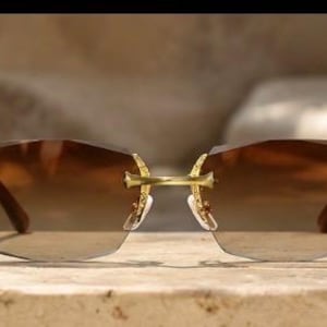 May include: Rimless sunglasses featuring brown gradient lenses and a gold-tone metal frame. The temples and bridge display intricate engraved detailing. The lenses are rectangular with slightly rounded edges, connected by a thin gold bridge.