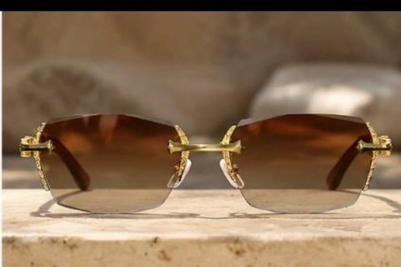 May include: Rimless sunglasses featuring brown gradient lenses and a gold-tone metal frame. The temples and bridge display intricate engraved detailing. The lenses are rectangular with slightly rounded edges, connected by a thin gold bridge.