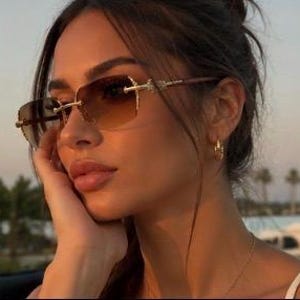 May include: Rimless sunglasses featuring gradient brown lenses that transition from dark to light. The frames have gold-toned metal accents at the hinges and bridge, with thin, sleek arms that extend along the sides of the face.