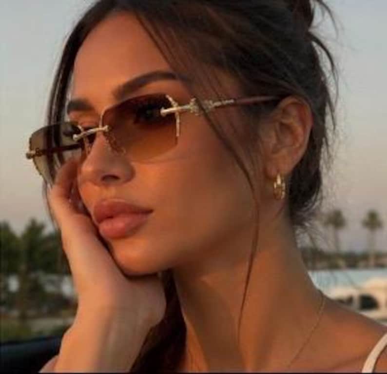 May include: Rimless sunglasses featuring gradient brown lenses that transition from dark to light. The frames have gold-toned metal accents at the hinges and bridge, with thin, sleek arms that extend along the sides of the face.