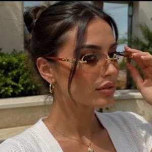 May include: Rimless sunglasses featuring gradient brown lenses and gold-tone metal temples with intricate detailing. The eyewear has a bridge design that connects the lenses, providing a sleek look. These glasses are worn with gold hoop earrings.