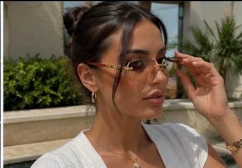 May include: Rimless sunglasses featuring gradient brown lenses and gold-tone metal temples with intricate detailing. The eyewear has a bridge design that connects the lenses, providing a sleek look. These glasses are worn with gold hoop earrings.