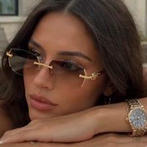 May include: Rimless sunglasses featuring gradient brown lenses and gold-tone metal temples with a cross-shaped emblem. A gold-tone metal wristwatch with a round face and a link bracelet is worn on the wrist, complementing the eyewear design.