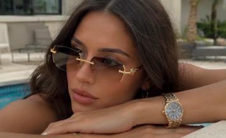 May include: Rimless sunglasses featuring gradient brown lenses and gold-tone metal temples with a cross-shaped emblem. A gold-tone metal wristwatch with a round face and a link bracelet is worn on the wrist, complementing the eyewear design.