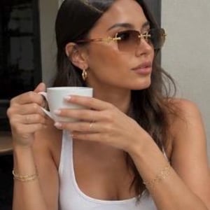 May include: Rimless sunglasses with gradient brown lenses and gold-tone metal temples, paired with a white tank top, gold hoop earrings, and two delicate gold chain bracelets. The eyewear features a rectangular shape with a thin bridge design.