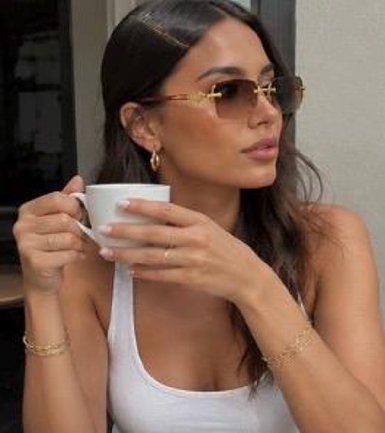 May include: Rimless sunglasses with gradient brown lenses and gold-tone metal temples, paired with a white tank top, gold hoop earrings, and two delicate gold chain bracelets. The eyewear features a rectangular shape with a thin bridge design.
