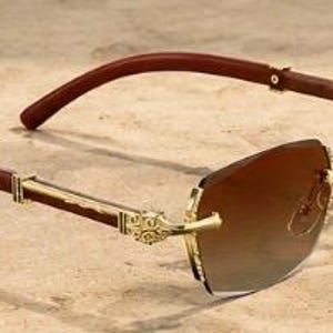 May include: Rimless sunglasses featuring gradient brown lenses and gold-tone metal temples with intricate engravings. The arms are constructed from dark brown wood, connecting to the frame with gold hinges, creating a distinct contrast.