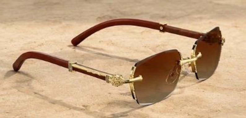 May include: Rimless sunglasses featuring gradient brown lenses and gold-tone metal temples with intricate engravings. The arms are constructed from dark brown wood, connecting to the frame with gold hinges, creating a distinct contrast.
