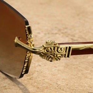 May include: Rimless sunglasses featuring brown tinted lenses and gold-tone metal temples. The hinges and temple arms are decorated with intricate embossed scrollwork patterns, and the bridge connection point displays a detailed metal design.