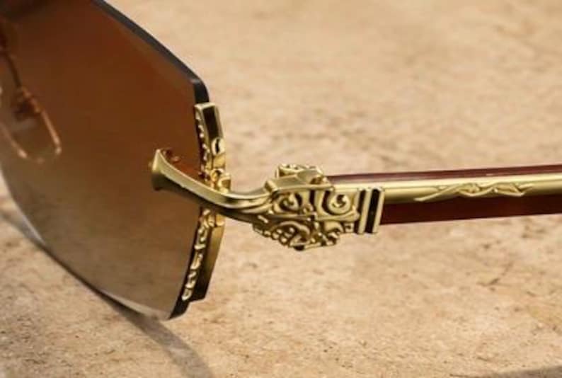 May include: Rimless sunglasses featuring brown tinted lenses and gold-tone metal temples. The hinges and temple arms are decorated with intricate embossed scrollwork patterns, and the bridge connection point displays a detailed metal design.