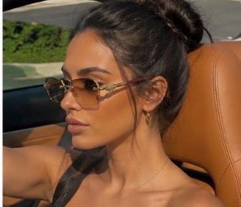 May include: Rimless sunglasses featuring a gradient brown lens that transitions from a darker shade at the top to a lighter tint at the bottom. The frames include gold-toned metal temples with intricate detailing and a bridge connecting the lenses.