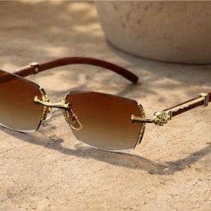 May include: Rimless sunglasses featuring gradient brown lenses that transition from dark to light. The frame includes gold-tone metal accents at the bridge and hinges, paired with dark brown temples that have a smooth, polished finish.