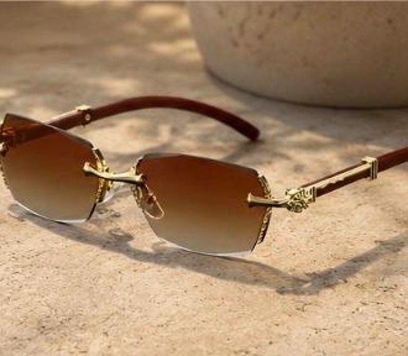 May include: Rimless sunglasses featuring gradient brown lenses that transition from dark to light. The frame includes gold-tone metal accents at the bridge and hinges, paired with dark brown temples that have a smooth, polished finish.