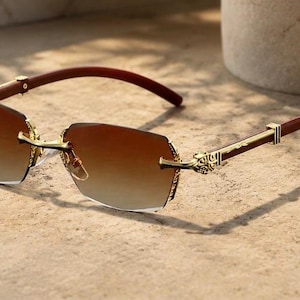 May include: Rimless sunglasses featuring gradient brown lenses and gold-tone metal hinges with intricate engraved detailing. The temples are constructed from a dark brown material, connecting to the gold-tone bridge and lens mounts.