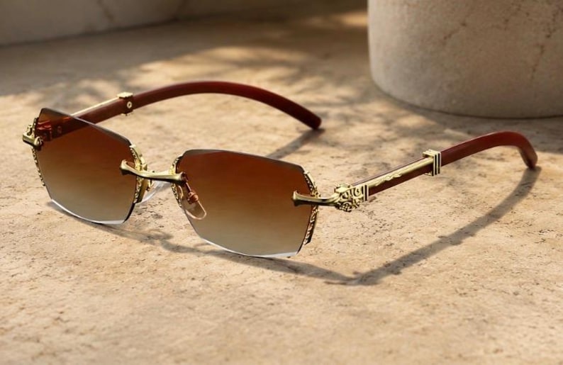 May include: Rimless sunglasses featuring gradient brown lenses and gold-tone metal hinges with intricate engraved detailing. The temples are constructed from a dark brown material, connecting to the gold-tone bridge and lens mounts.