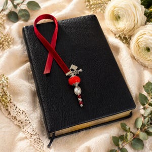 May include: A black leather-bound book featuring a red velvet ribbon bookmark. The bookmark is adorned with a silver-toned charm, a textured red spherical bead, a clear oval crystal, and a small red bead at the end of the dangling ornament.