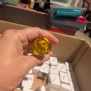 May include: A faceted, round, translucent yellow glass or acrylic knob or decorative sphere. The object is held above a cardboard box containing multiple small white paper boxes, some of which are labeled with handwritten text like Pink, Lime, and Red.