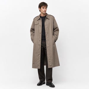 May include: A long, taupe-colored trench coat featuring a classic collar, shoulder epaulets, and a button-front closure. The coat is worn over a black sweater and dark brown trousers, paired with black leather dress shoes on a plain background.