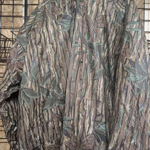 May include: A hooded rain jacket featuring a Realtree camouflage pattern with brown, green, and grey bark and leaf motifs. The jacket includes a front button closure, a drawstring hood, and long sleeves, with the Realtree brand name printed on the front.
