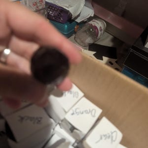 Può includere: Una collezione di piccole scatole quadrate bianche disposte in un contenitore di cartone, ognuna etichettata con testo scritto a mano che include Black, Orange, Clear e Red, con un oggetto circolare scuro sfocato tenuto in primo piano.