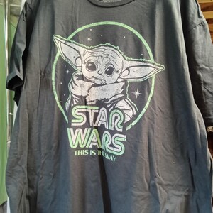 May include: A charcoal gray short-sleeve t-shirt featuring a graphic of Grogu from Star Wars inside a light green circle. The design includes the text Star Wars and This Is The Way in light green lettering with a distressed, weathered appearance.