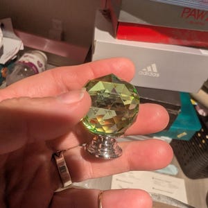 Può includere: Un pomello per mobile in cristallo verde lime sfaccettato con una base in metallo color argento lucido. Il pomello presenta un design geometrico a molteplici sfaccettature che cattura la luce, fissato su un supporto circolare in metallo argentato a gradini.