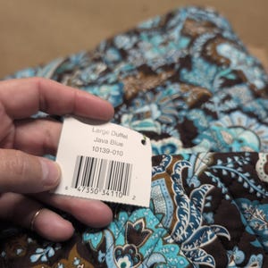 May include: A quilted fabric duffel bag featuring a dark brown, light blue, and white paisley pattern. A white rectangular tag is attached, displaying the text Large Duffel, Java Blue, 10139-010, and a barcode with the number 6 47350 34110 2.