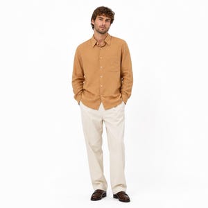 May include: A long-sleeved button-down shirt in a solid tan color featuring a single chest pocket and a classic collar, paired with straight-leg off-white trousers and dark brown leather dress shoes, creating a neutral-toned ensemble.