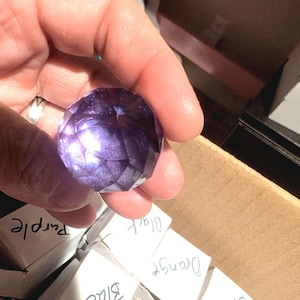 May include: A faceted, round, purple gemstone-shaped object with a shimmering, glittery interior texture. The object is held against a background of small white cardboard boxes, some of which are labeled with handwritten text including Purple, Black, Orange, and Clear.