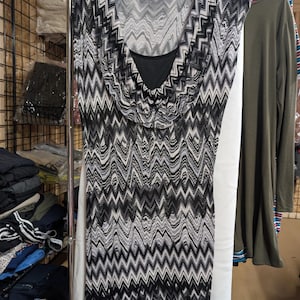Könnte beinhalten: Ein ärmelloses Kleid mit einem Zickzack-Chevron-Muster in Schwarz, Weiß und Grau. Das Kleidungsstück hat einen drapierten Wasserfall-Ausschnitt mit einer einfarbigen schwarzen Innenschicht im Brustbereich. Es hängt an einem Kleiderbügel mit Etikett.