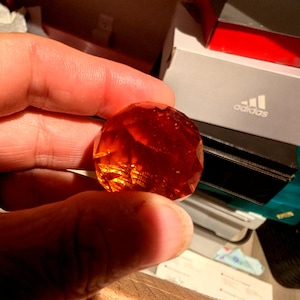 May include: A faceted, spherical object made of translucent, deep amber-colored glass. The surface features multiple geometric cuts that catch and reflect light, creating a glowing, warm orange appearance with visible internal clarity and depth.