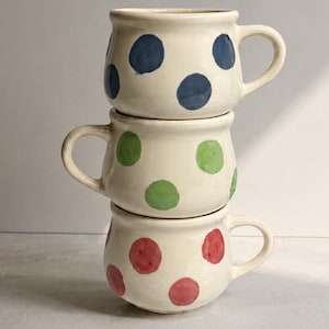 Può includere: Una pila di tre tazze in ceramica color crema con corpo arrotondato e manici. La tazza superiore presenta pois blu, la tazza centrale ha pois verdi e la tazza inferiore è decorata con pois rossi dipinti a mano.