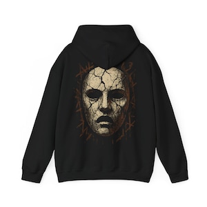 May include: A black hooded sweatshirt featuring a large graphic on the back. The design displays a beige, cracked human face mask with deep, dark eye sockets and a mouth, surrounded by a border of dark brown, jagged, linear markings.