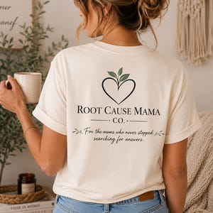 Può includere: Una t-shirt a maniche corte color crema con una grafica a forma di cuore nero sormontata da foglie verdi. Il testo recita: ROOT CAUSE MAMA CO. For the moms who never stopped searching for answers. La maglietta è abbinata a jeans blu.