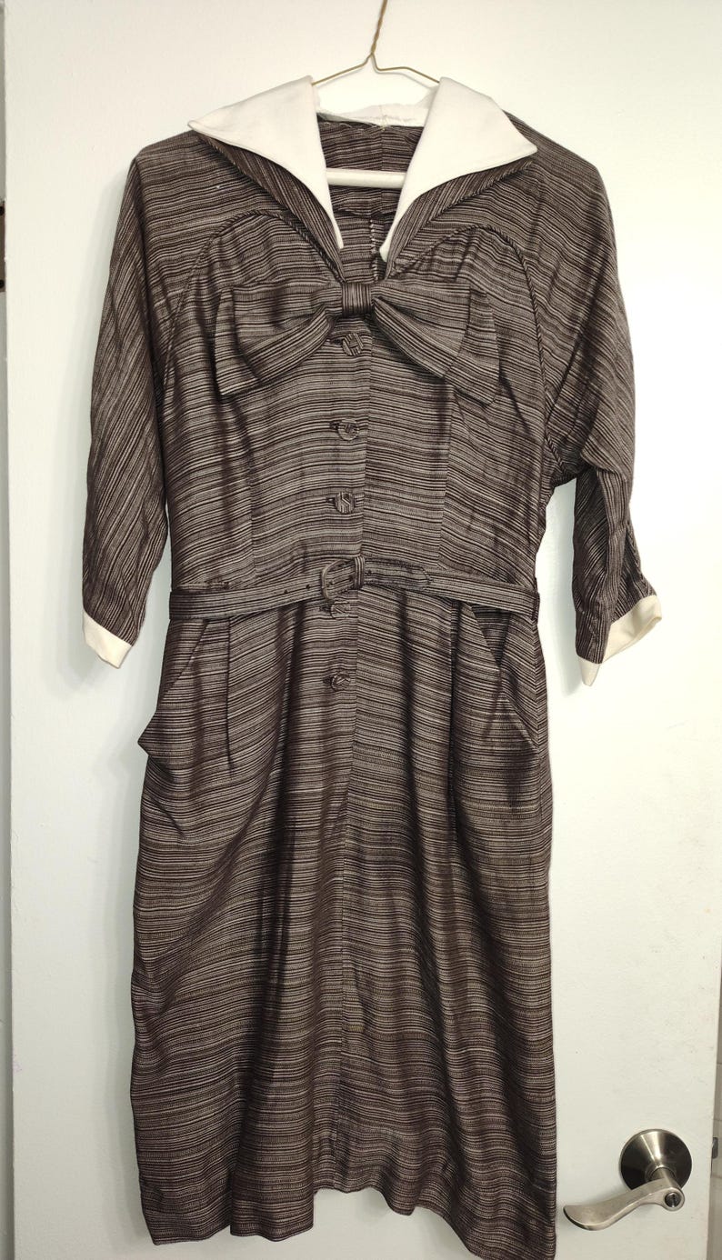 May include: A brown and white striped dress with a white collar and a bow at the neckline. The dress has a belt and pockets.