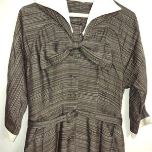 May include: A brown and white striped dress with a white collar and a large bow at the neckline. The dress has a belt and pockets.
