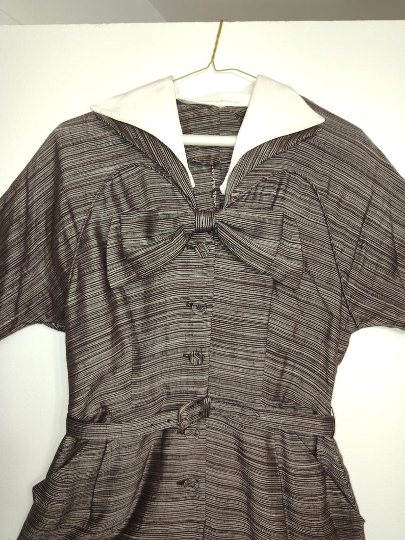 May include: A brown and white striped dress with a white collar and a bow at the neckline. The dress has a belt and short sleeves.