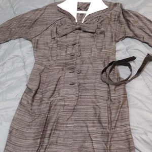 May include: A brown and white striped dress with a white collar and a bow at the neckline. The dress has a button-up front and a black belt.