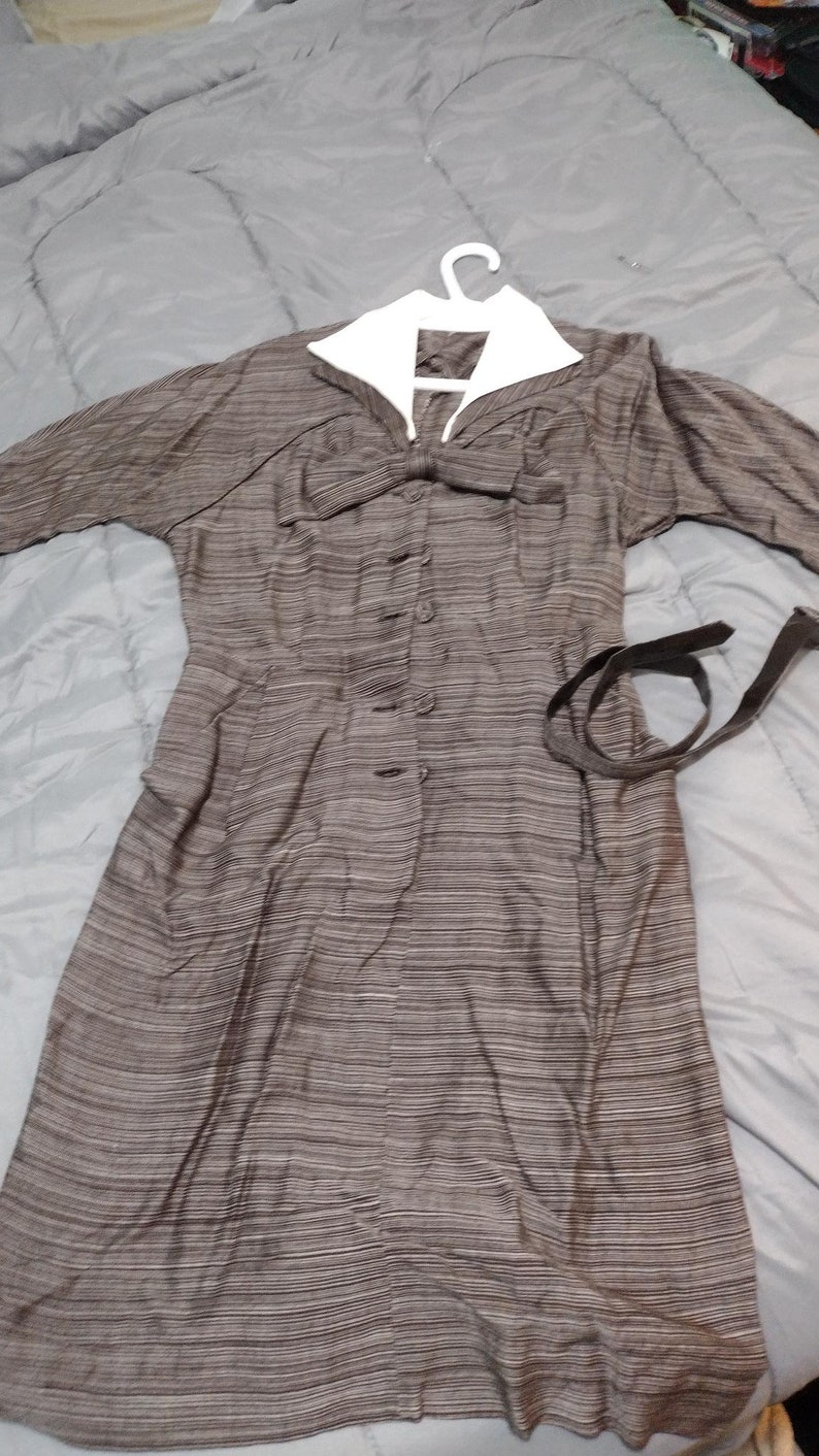 May include: A brown and white striped dress with a white collar and a bow at the neckline. The dress has a button-up front and a black belt.