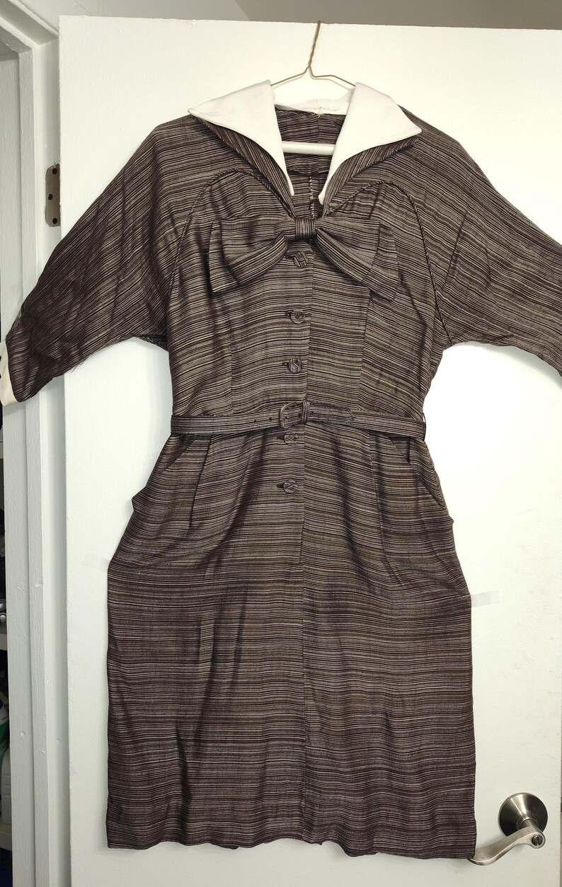 May include: A brown and white striped dress with a white sailor collar and a bow at the neckline. The dress has a belt and buttons down the front.