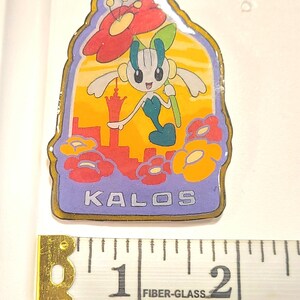 May include: A rectangular enamel pin featuring a cartoon character holding a large red flower against a yellow and orange sunset background with a red city silhouette. The word KALOS is printed in white at the bottom. A measuring tape shows it is 2 inches wide.