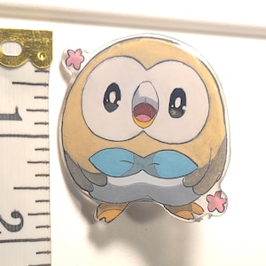 May include: A round, tan and white acrylic pin featuring a cartoon owl with large black eyes, a light blue bowtie, and small orange feet. Two small pink cherry blossom shapes are attached to the sides. A white measuring tape shows a height of 5.08 cm.