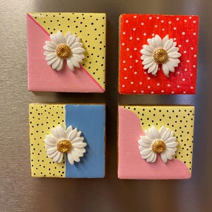 May include: Four square magnets featuring hand-painted designs with yellow, pink, red, and blue sections, black polka dots, and a three-dimensional white daisy with a textured gold centre attached to the front of each individual piece.