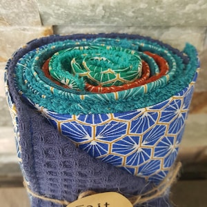 Peut inclure: Un ensemble de tissus roulés aux motifs géométriques bleus, verts et orange avec des surfaces texturées. Une petite étiquette ovale en bois attachée avec de la ficelle est fixée à l'avant, affichant le texte Fait main avec un cœur rouge.