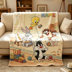 May include: A cream-coloured fleece blanket featuring a colourful illustration of Looney Tunes characters including Bugs Bunny painting, Tweety Bird holding a spool, the Tasmanian Devil spinning, and Sylvester the Cat playing with yarn, with the text NAME.