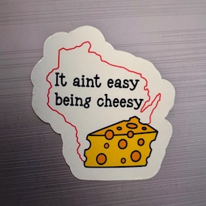 May include: A die-cut sticker featuring a red outline of the state of Wisconsin with the black text It aint easy being cheesy printed across the center and a yellow wedge of Swiss cheese with orange holes positioned at the bottom right corner.